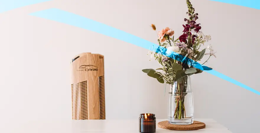 Květina ve váze na bílém stole, v pozadí čistička vzduchu Ionic-CARE v dekoru dřevo dub